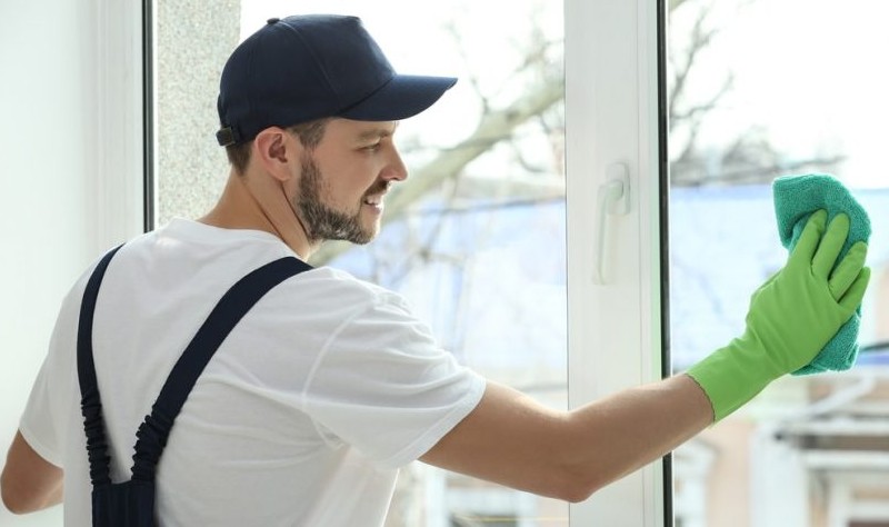 a young man wearing a blue cap and green gloves wiping window glass with a rag