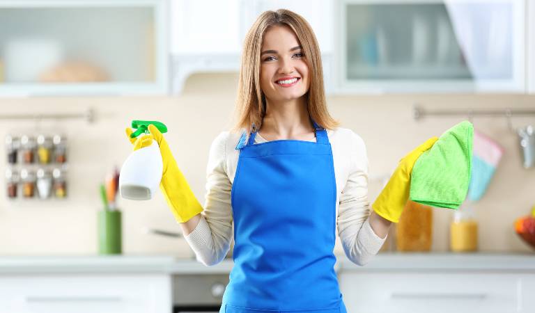 Woman in white blue dress and yellow gloves holding white bottle in one hand and green cloth in second hand