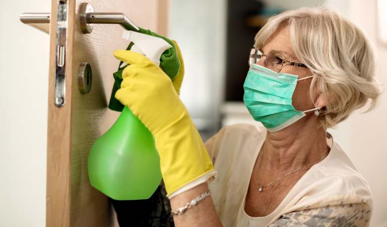 Old woman in mask and yellow glove holding a green bottle cleaning door.