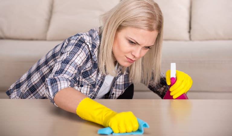 Woman in check shirt and yellow gloves holding a red bottle in her hand and scrubbing table with green cloth.