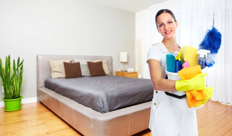 Woman in white dress and yellow gloves holding a basket filled with tools and supplies inside a bedroom.