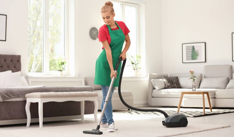 Woman in red-green dress vacuuming carpet inside living room.