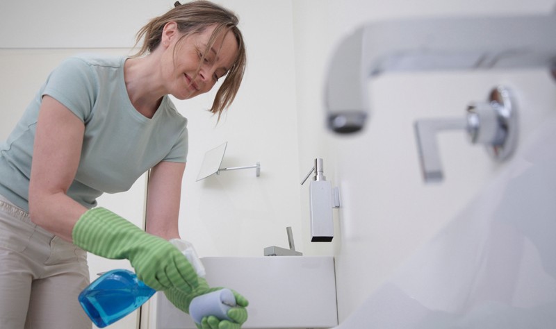 smiling young woman holding a spray bottle and a sponge inside a bathroom