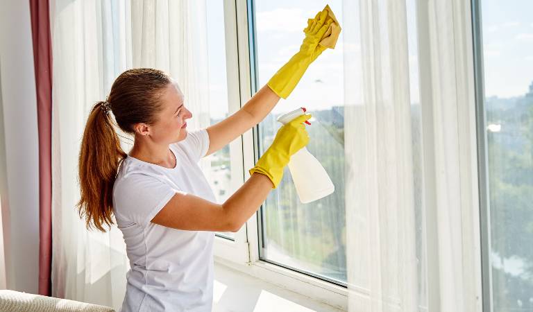 Woman in white top and yellow gloves holding a white bottle cleaning curtains.