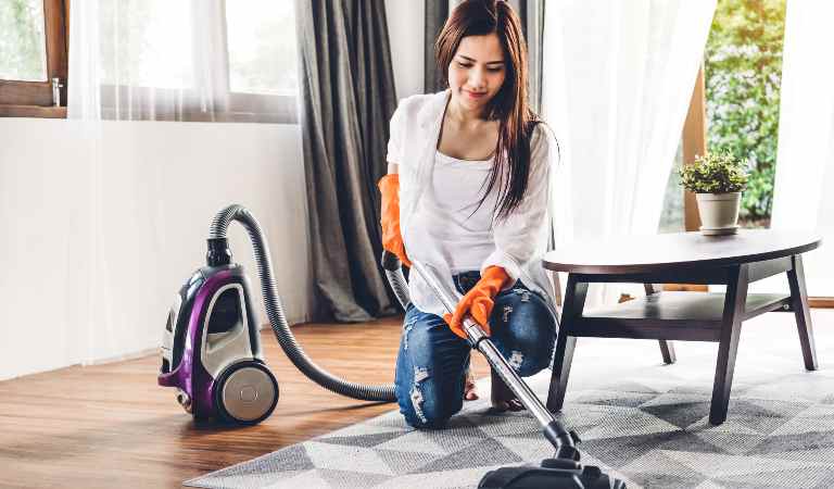Woman in white top, blue jeans and orange gloves vacuuming carpet inside living room