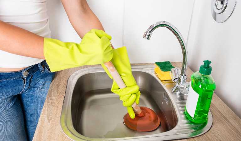 Woman in white top and blue jeans cleaning drain inside her kitchen.