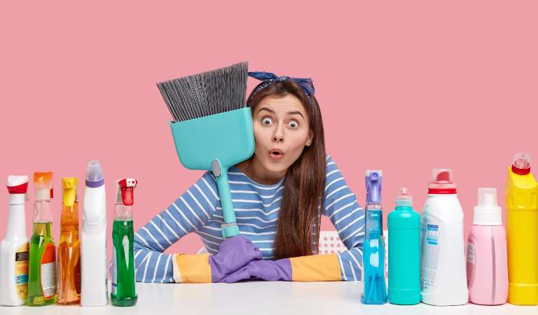 Woman holding a big brush and sitting with different bottels and cleaning tools.