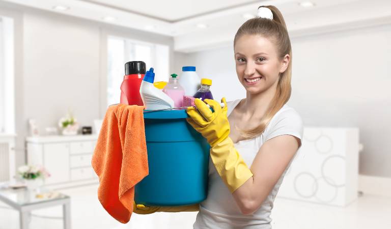 Woman in white top holding a blue basket filled with tools, cloth and bottles