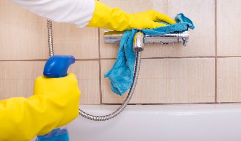 Woman in yellow gloves holding a blue bottle in her hand cleaning shower with a cloth.