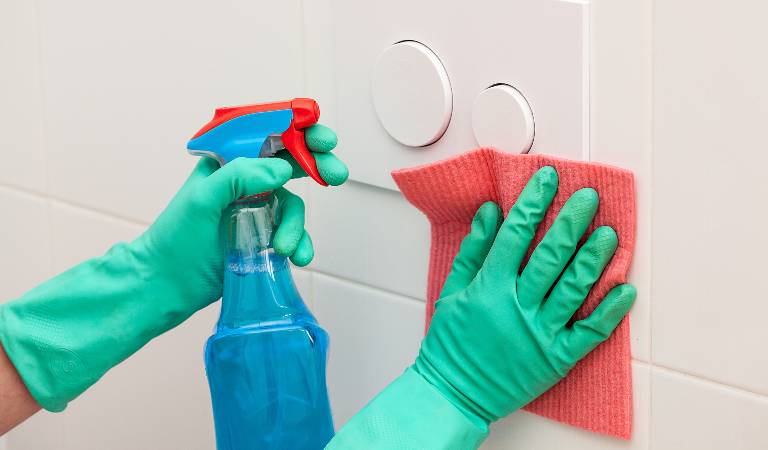 Hand of a woman in green gloves holding a blue bottle cleaning tiles with red cloth.