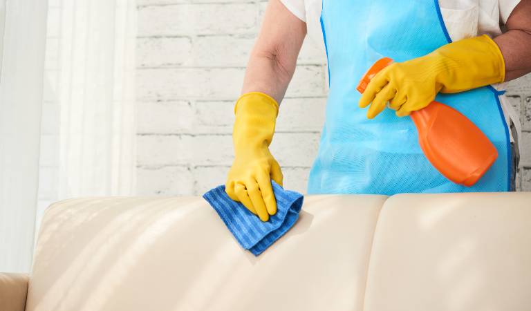 Cleaner in white-blue uniform and yellow gloves holding a orange bottle and cleaning Upholstery Fabrics with blue cloth.
