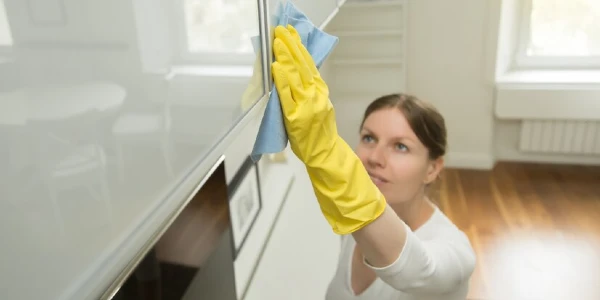 A lady in white top yellow gloves cleaning cabinets in kitchen.