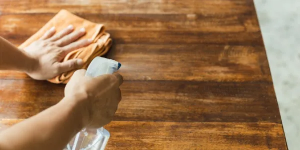 man holding a bottle in hand cleaning wooden table with a cloth.