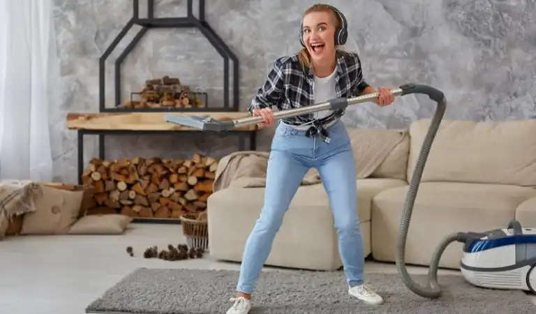 Lady in check shirt and blue jeans with headphone holding a vacuum cleaner.